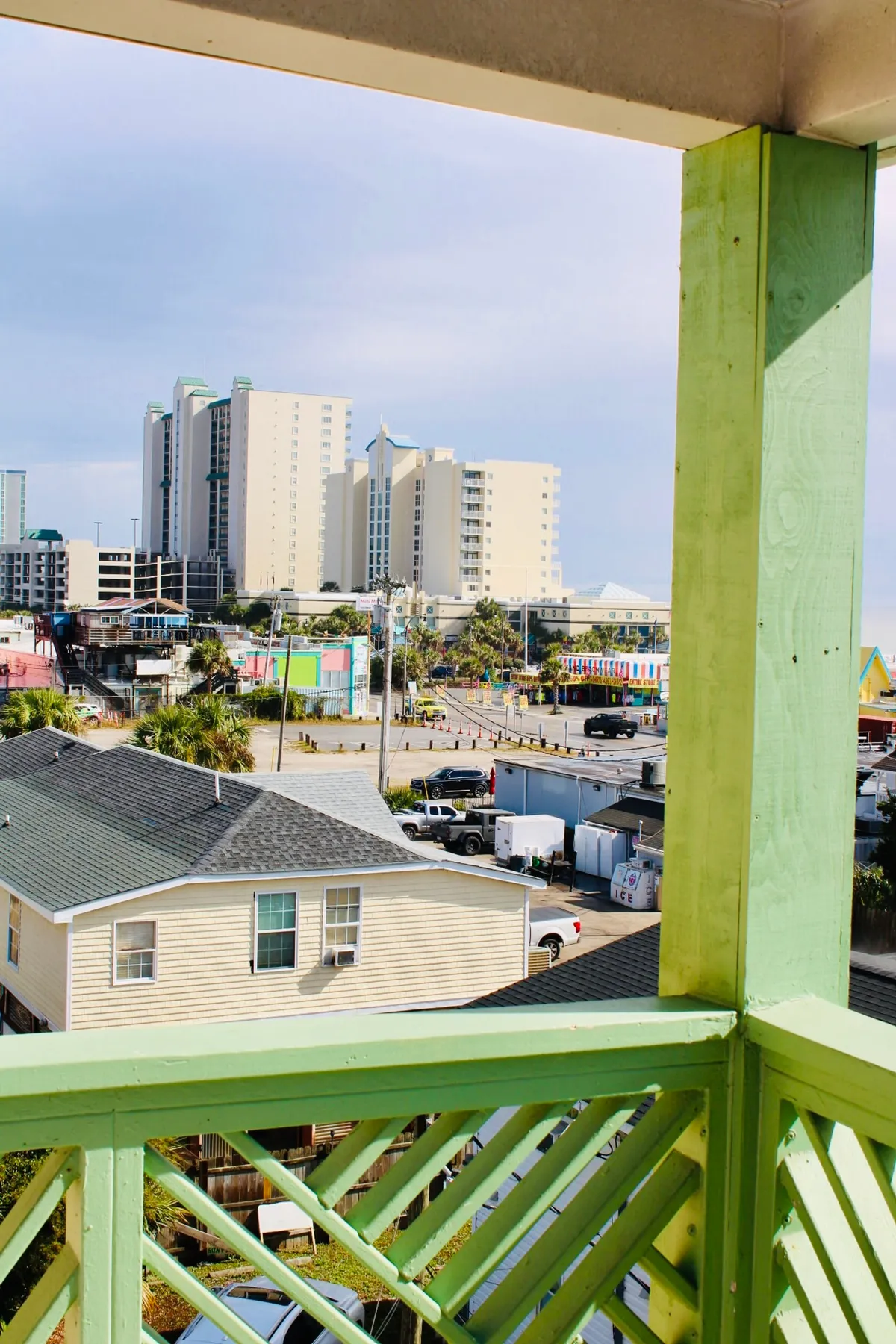 From a vantage point, this view captures colorful buildings and tall hotels along the beachfront, with a balcony railing in the foreground. The lively atmosphere suggests people-watching, local festivities, and the pleasure of enjoying the ocean from a private perch, especially during mornings or at sunset.
