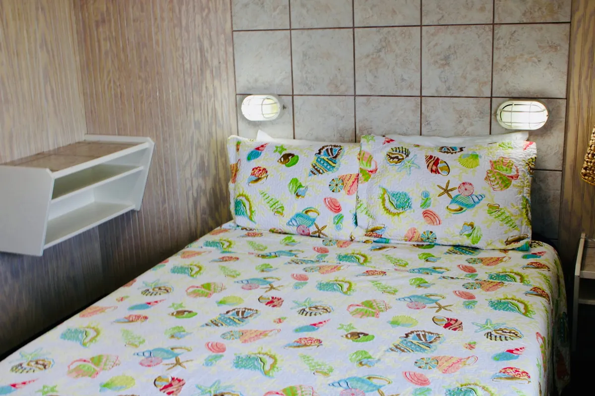 A sunlit bed topped with a colorful bedspread and decorative pillows, framed by wall-mounted lights. Natural light pours in, accentuating the relaxed, casual coastal décor and creating a cozy atmosphere for unwinding.