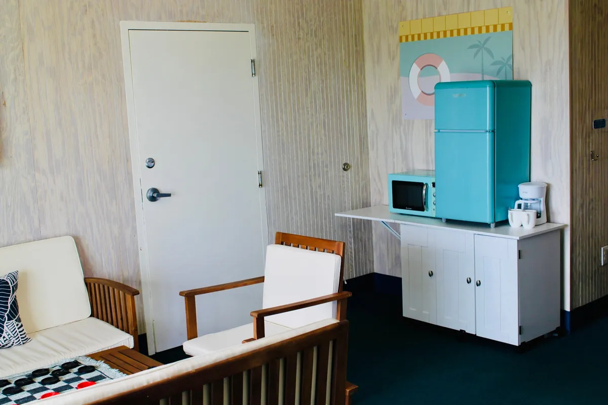 An inviting living area featuring an aqua refrigerator, white sofa, wooden chairs, and colorful wall art. The nostalgic, cheerful decor encourages relaxation, storytelling, and gathering with friends or family.