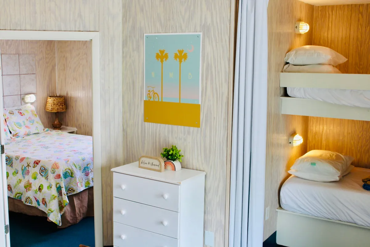 A colorful, cheerful open-plan space with a main bed featuring a vibrant bedspread, adjacent bunk beds for extra guests, framed art on the walls, and a dresser. The atmosphere feels inviting, perfect for family bonding or relaxing after a coastal adventure.